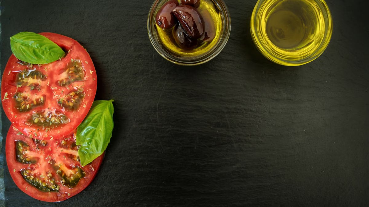 Top view of fresh basil, sliced tomatoes, olives, and olive oil on slate, perfect for Italian cuisine themes.